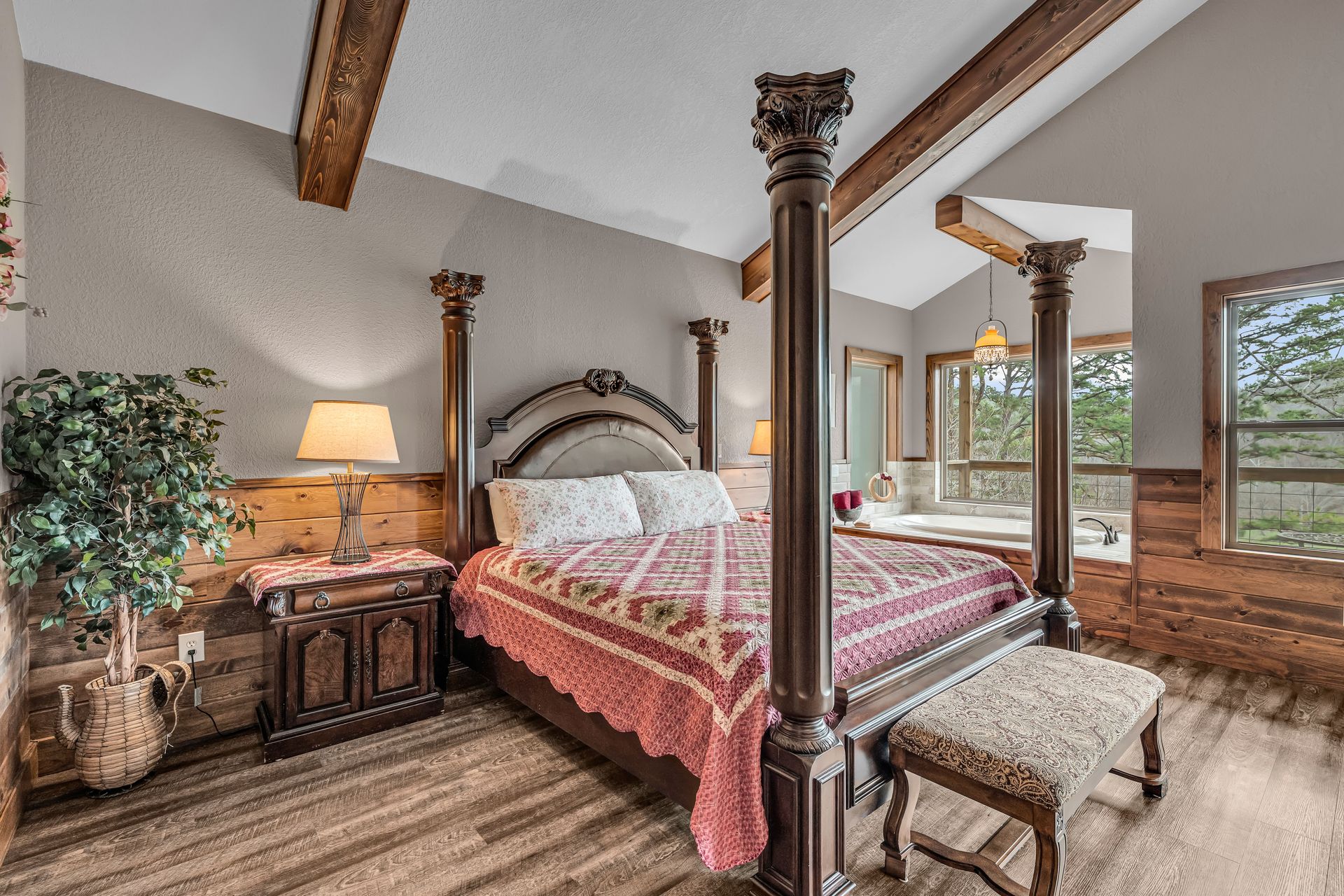 Bedroom with four-poster bed, wood beams, bedside lamp, and large windows overlooking greenery