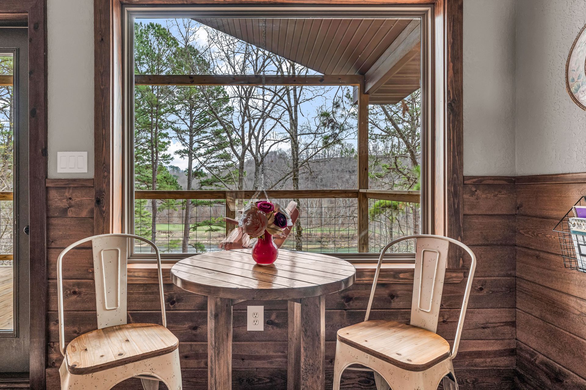 Small dining nook with round table, two chairs, and large window overlooking trees and a covered deck.