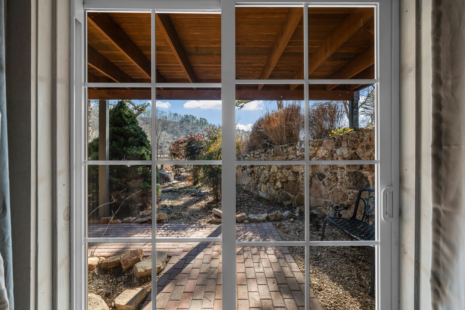 View through a white-paned window to a brick patio, trees, and hills under a wooden roof.