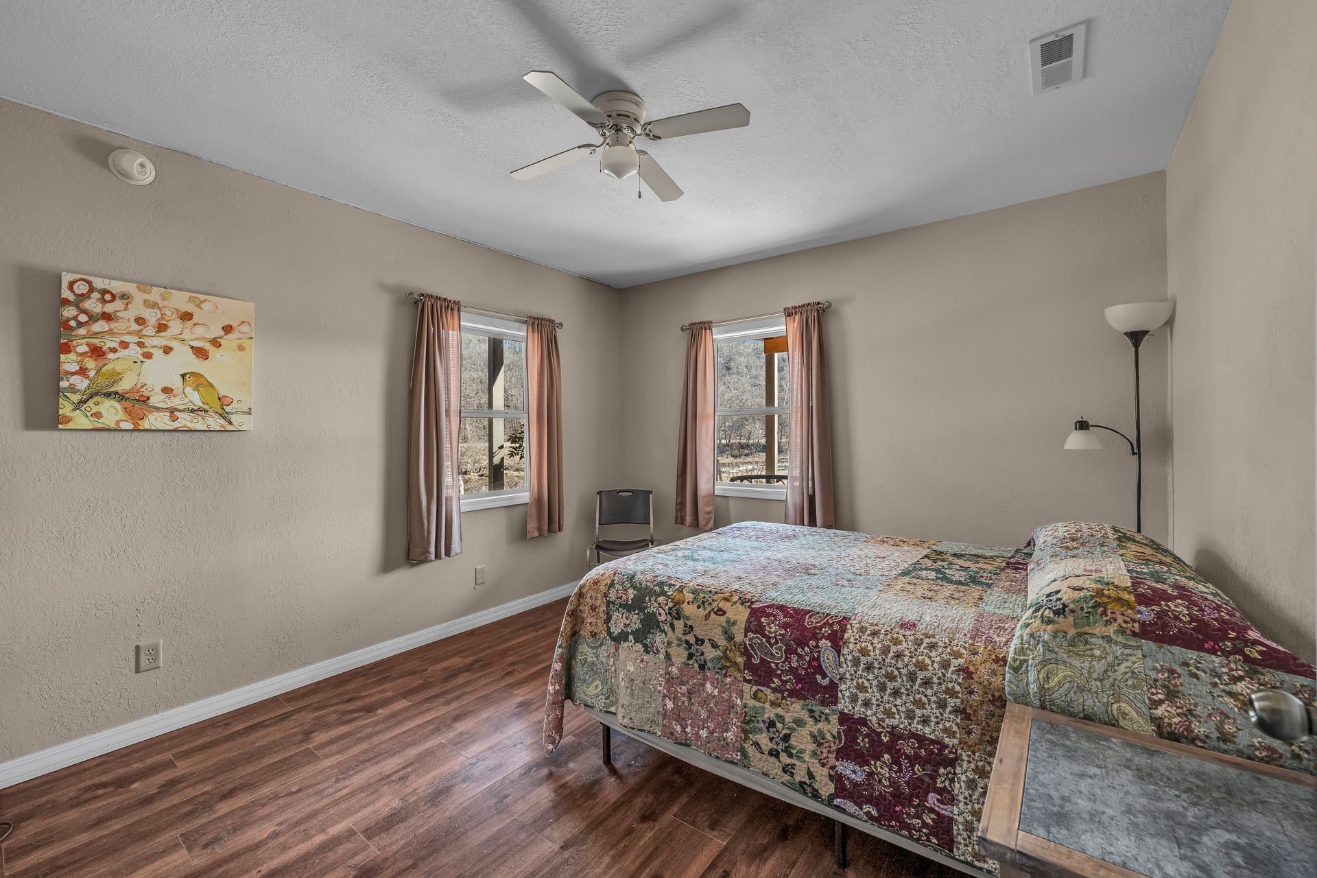 Bedroom with bed, two windows, wood floors, ceiling fan, and floor lamp.