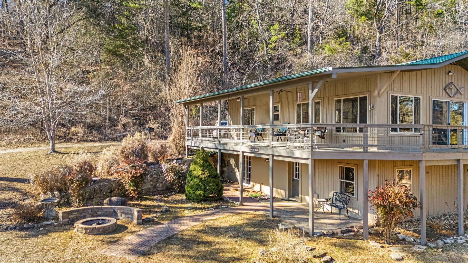 Two-story beige house with a covered porch beside a wooded hillside, viewed from the yard
