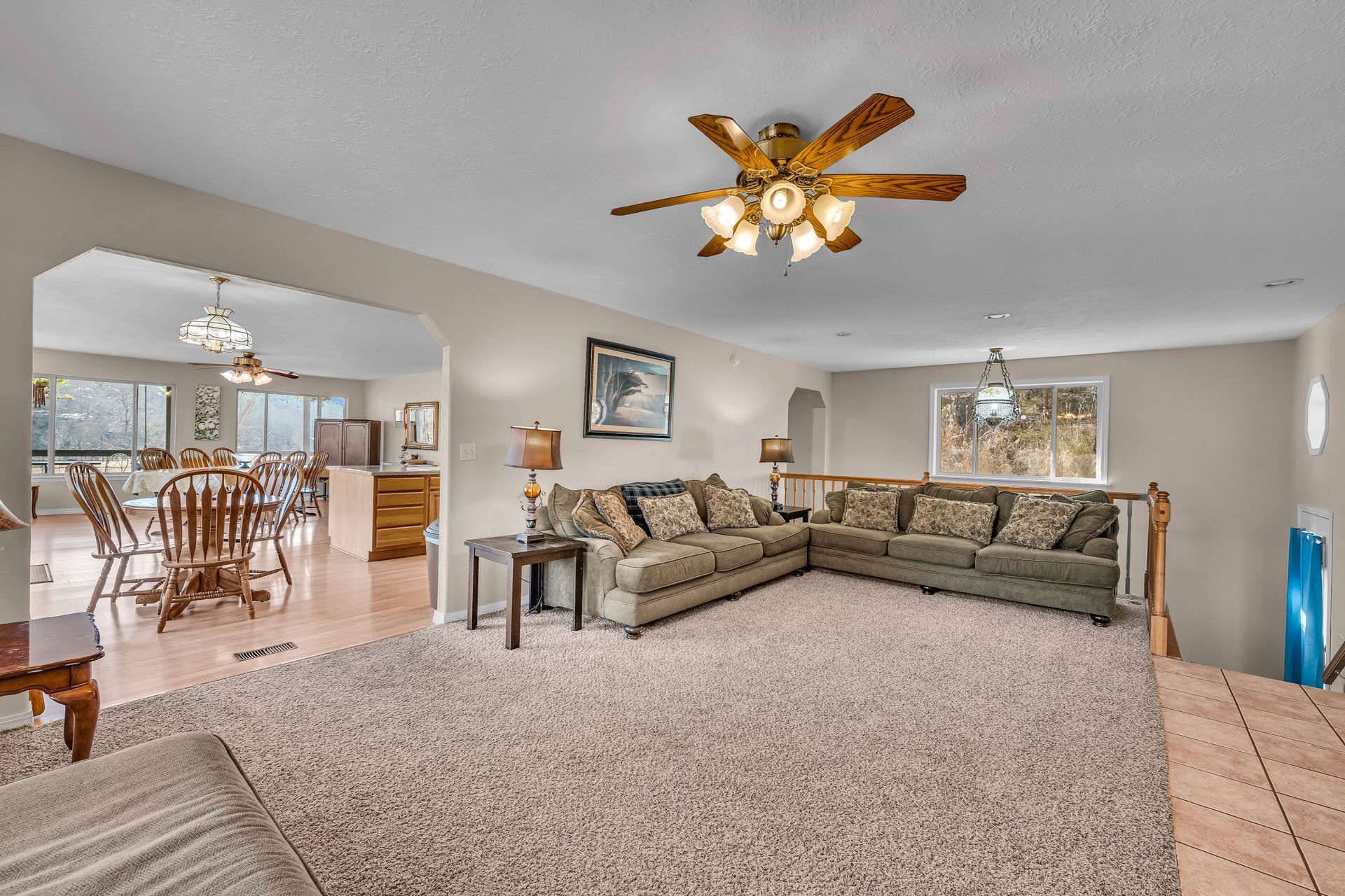 Spacious beige living room with sectional sofa, carpet, ceiling fan, and view into dining and kitchen area