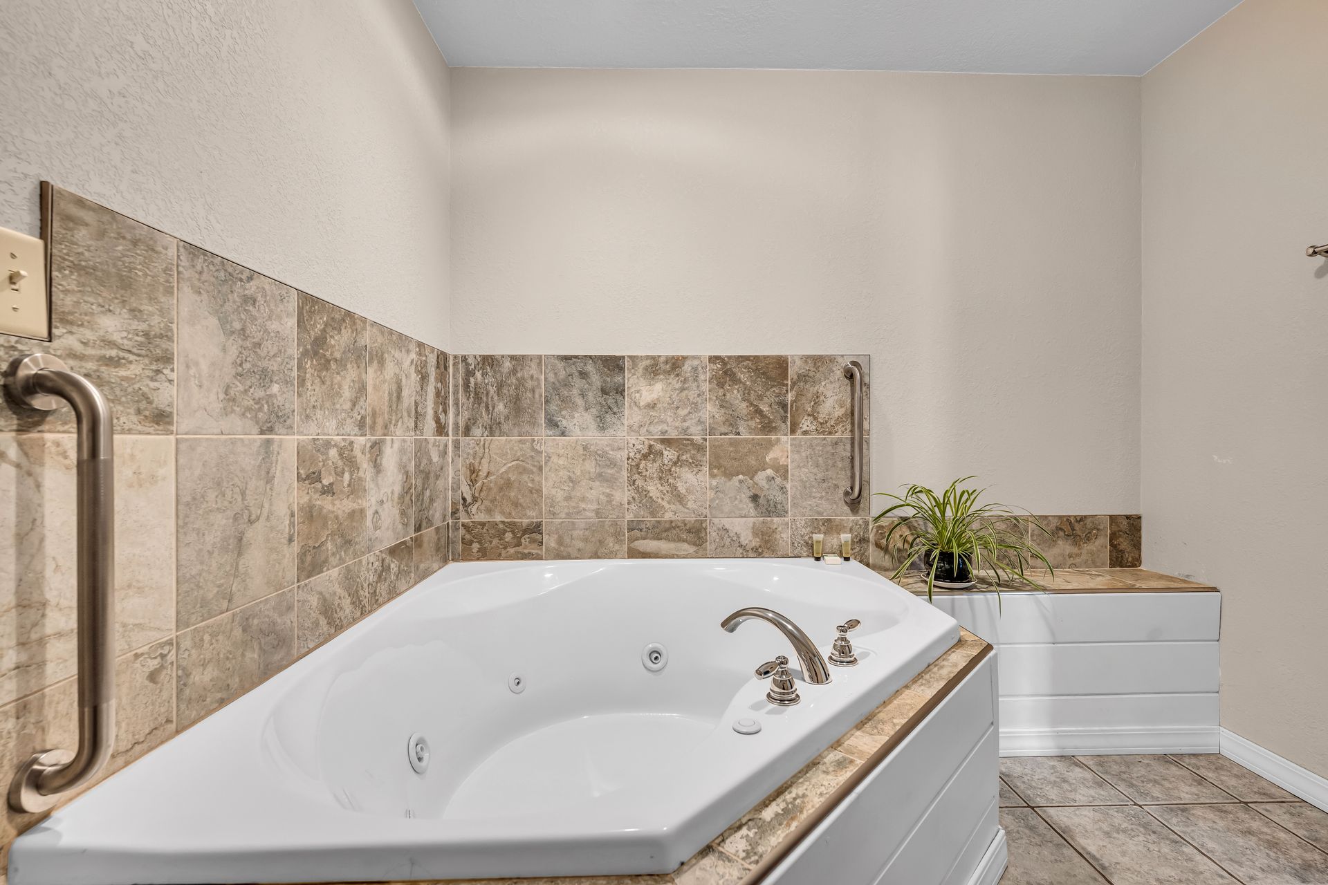White soaking tub in a tiled bathroom with beige stone walls and a chrome grab bar