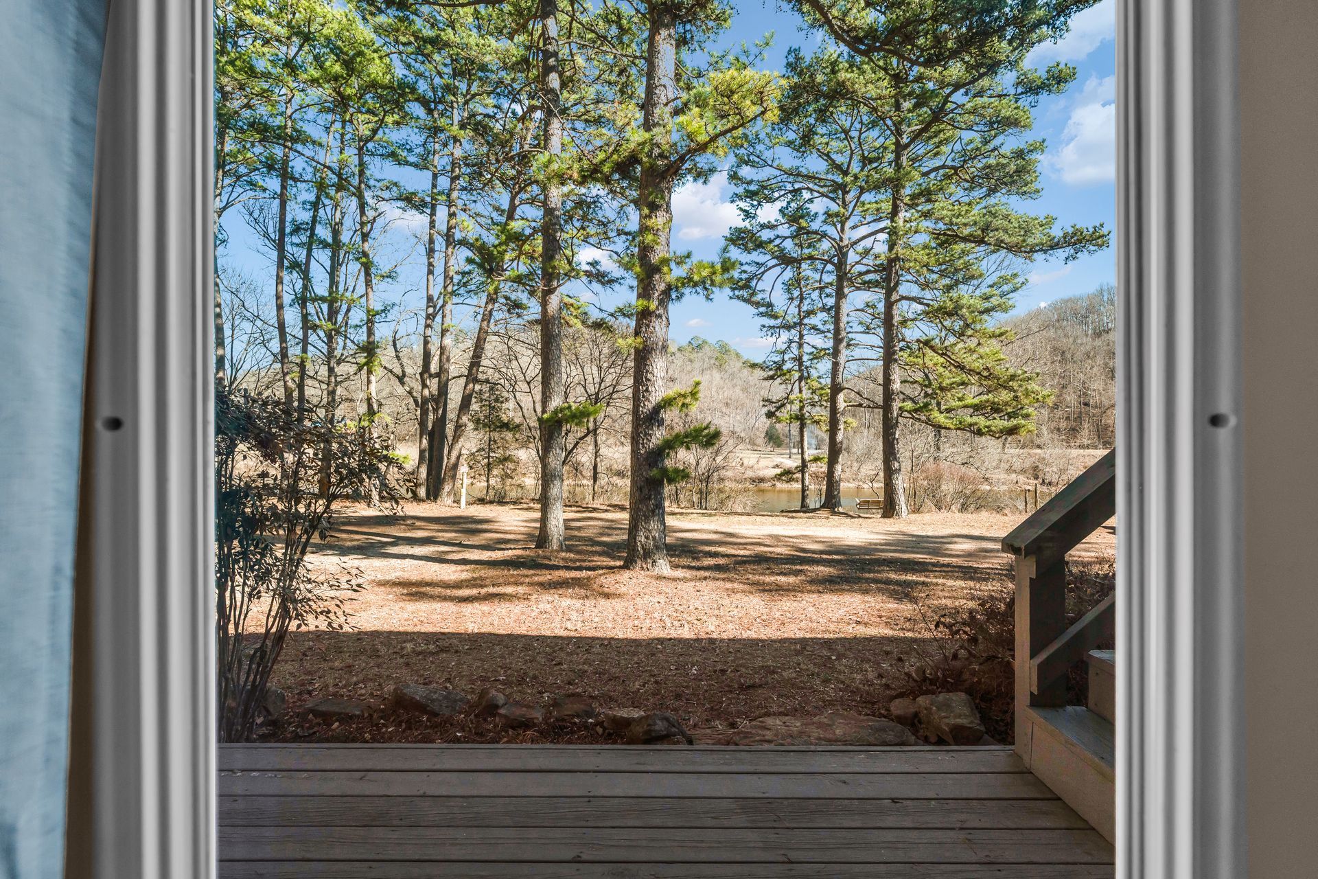 View through open doorway to pine trees, rocky ground, and blue sky beyond a wooden deck