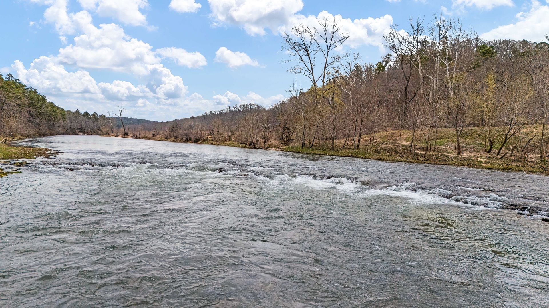 Wide river flowing through a rocky valley with leafless trees under a cloudy blue sky