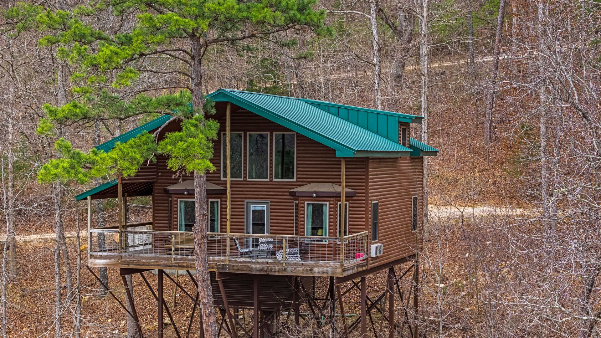 Rustic cabin on stilts in a leafless forest, with a green roof and wooden deck.