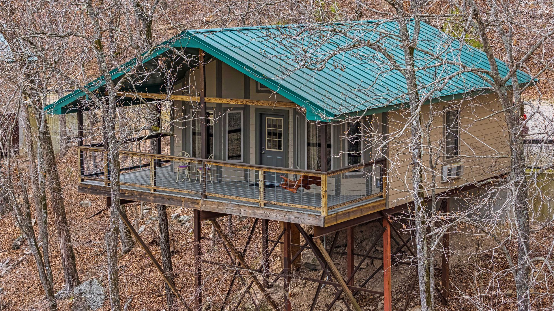 Cabin on stilts with a teal roof nestled among bare trees on a hillside