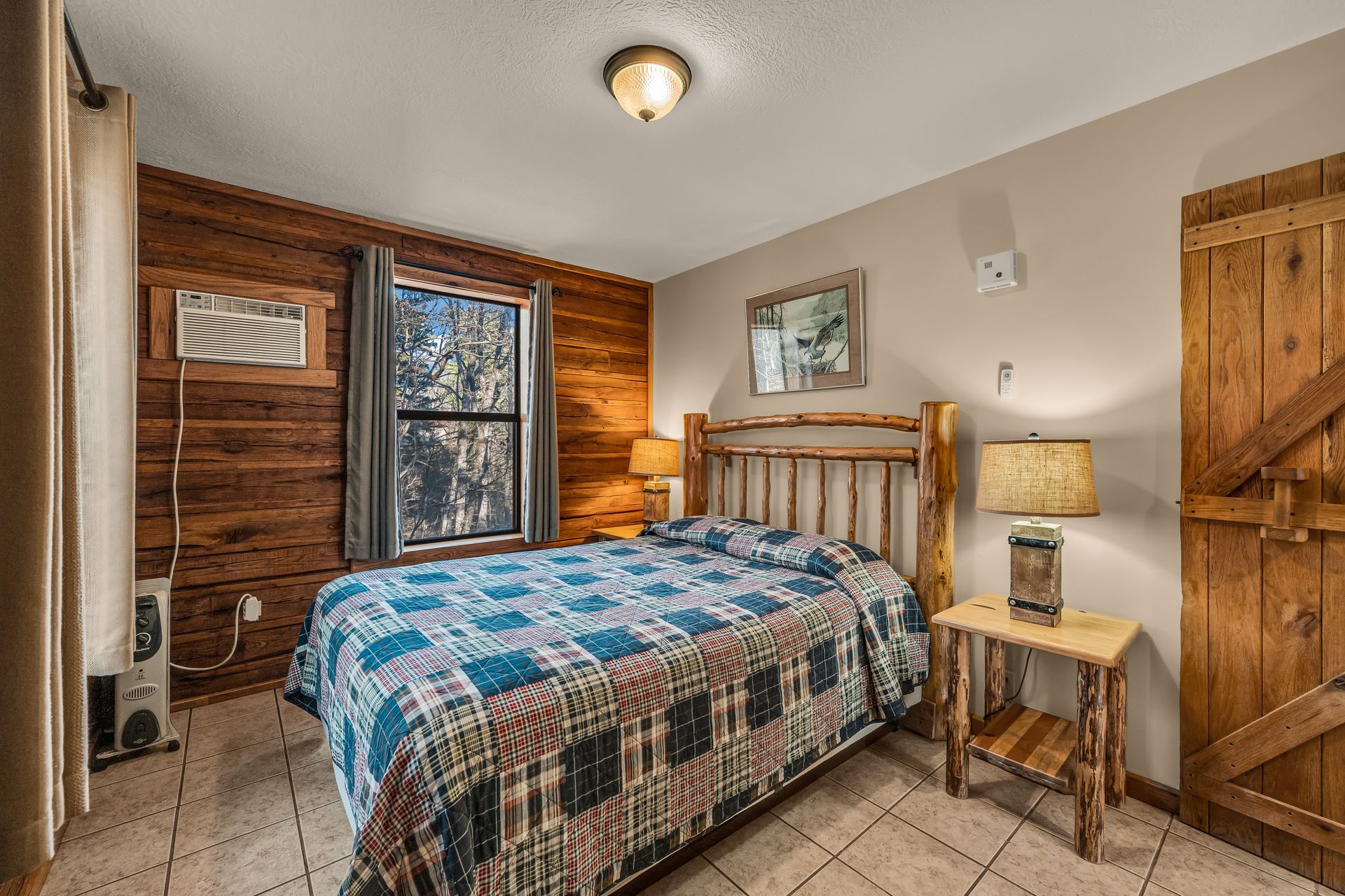 Small bedroom with a wooden bed, blue quilt, bedside lamp, window, and rustic wood shutters.