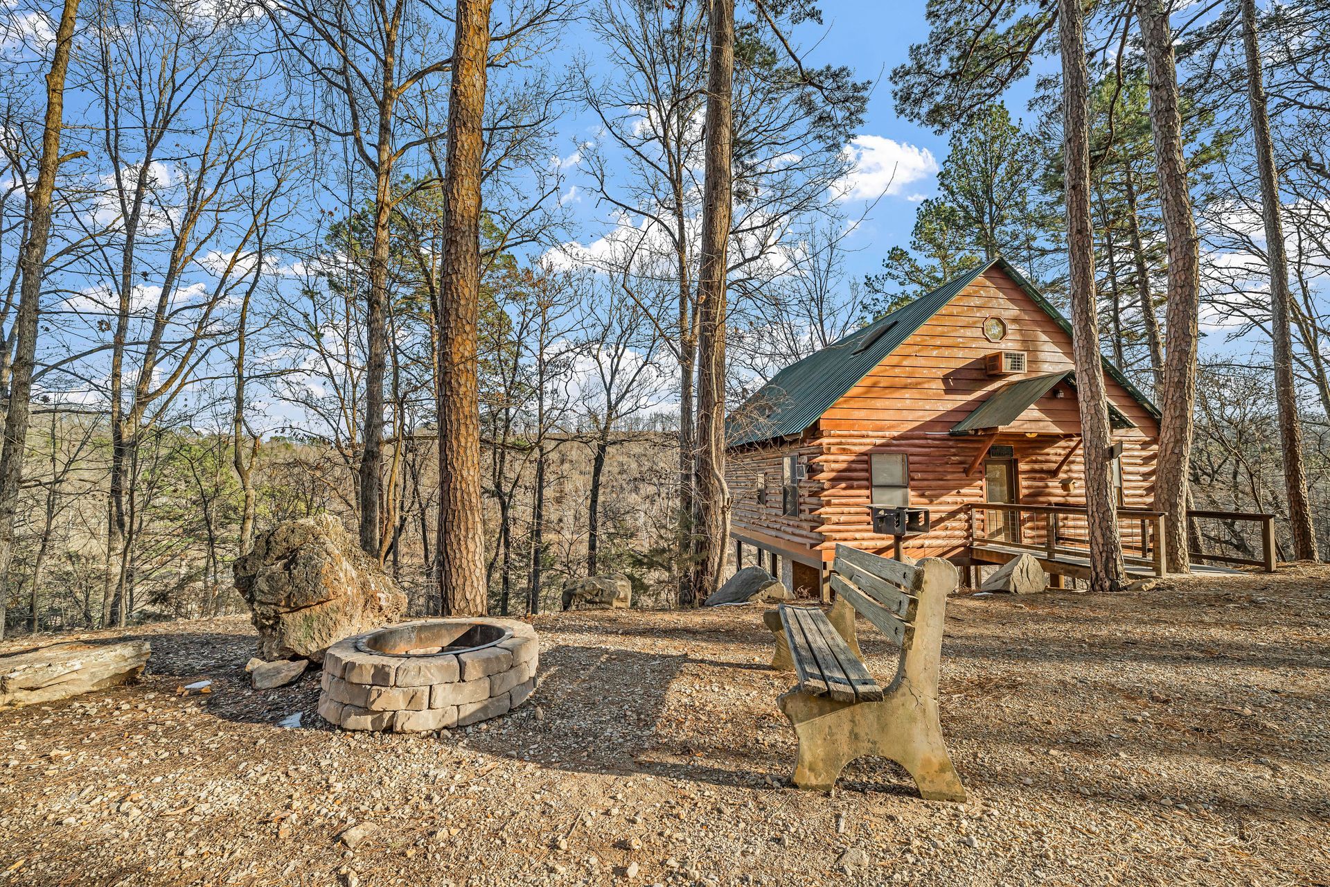 Rustic cabin in a wooded clearing with a fire pit and a wooden bench under a blue sky