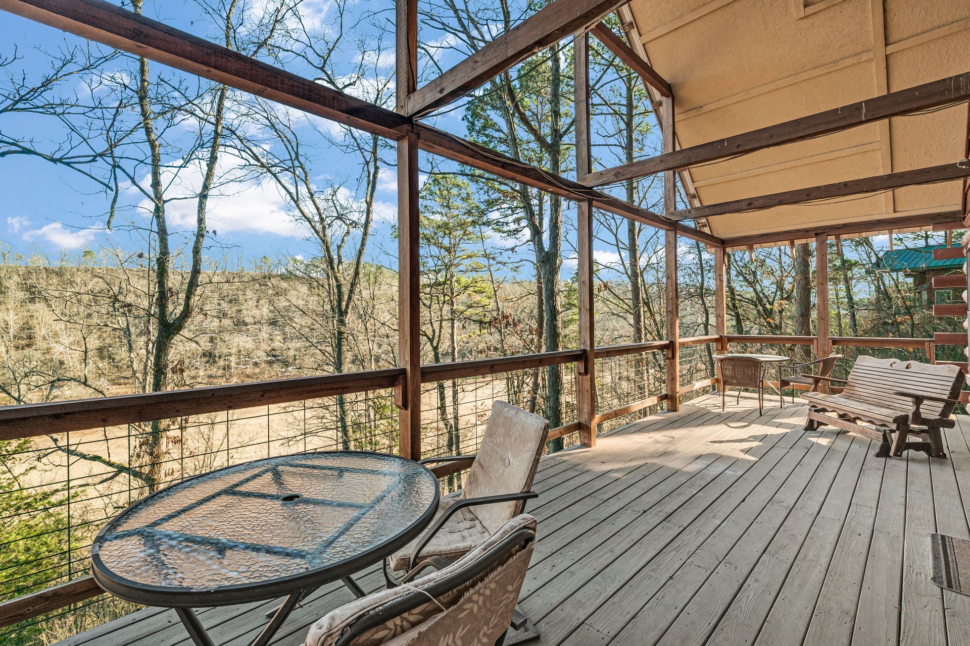 Covered wooden deck with glass table and chairs overlooking a wooded landscape and blue sky