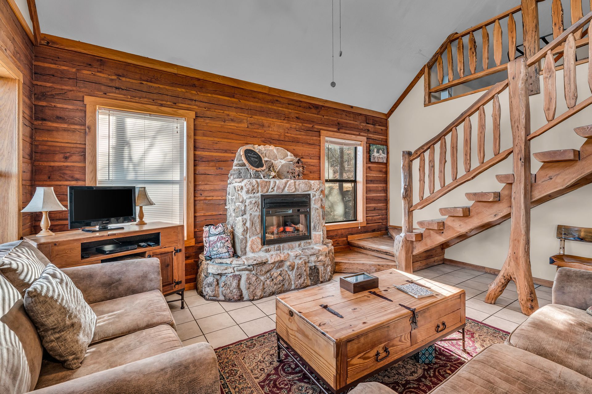 Cozy living room with stone fireplace, wooden staircase, beige couches, and TV near a window