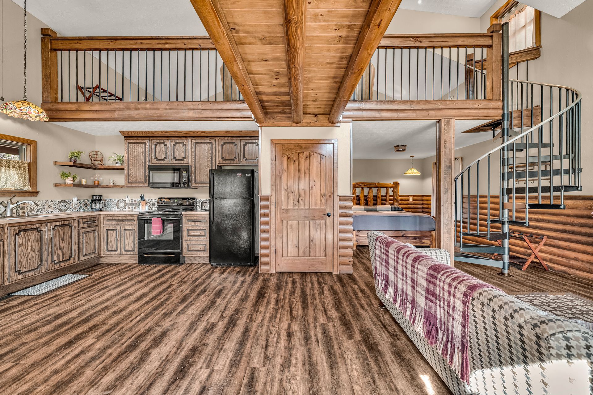 Rustic open-plan cabin interior with kitchen, loft, spiral staircase, and wood-paneled walls