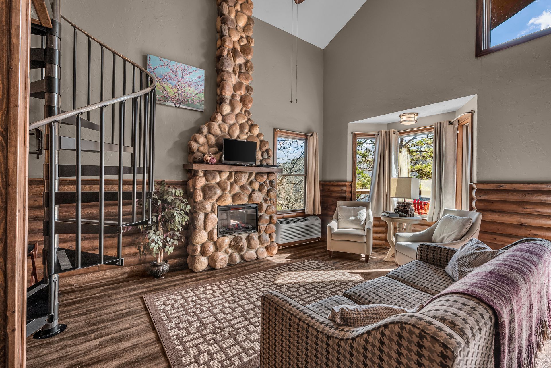 Cozy living room with stone fireplace, spiral staircase, and patterned sofa by a bright window.