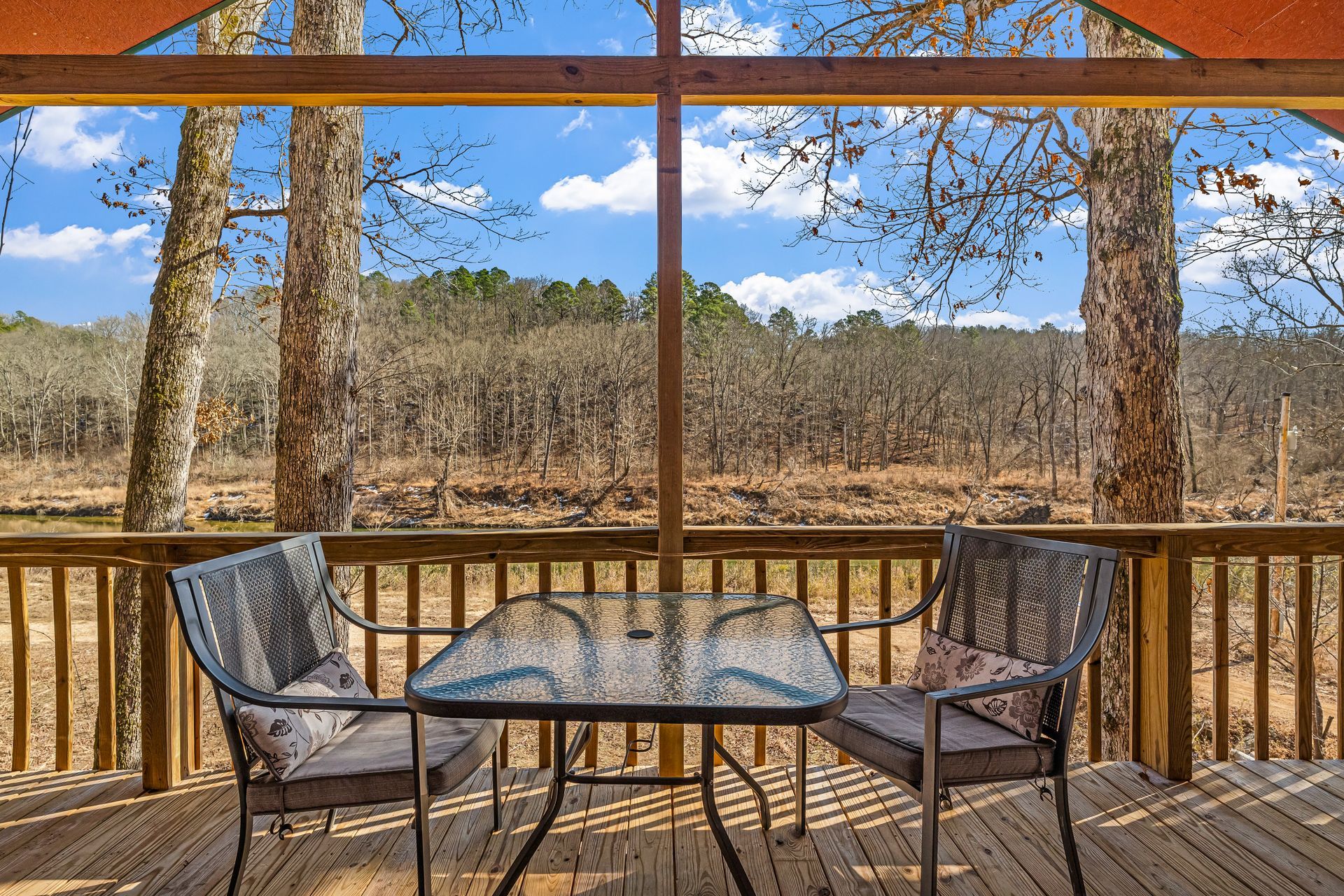 Wooden deck with glass table and two chairs overlooking a wooded landscape under a blue sky