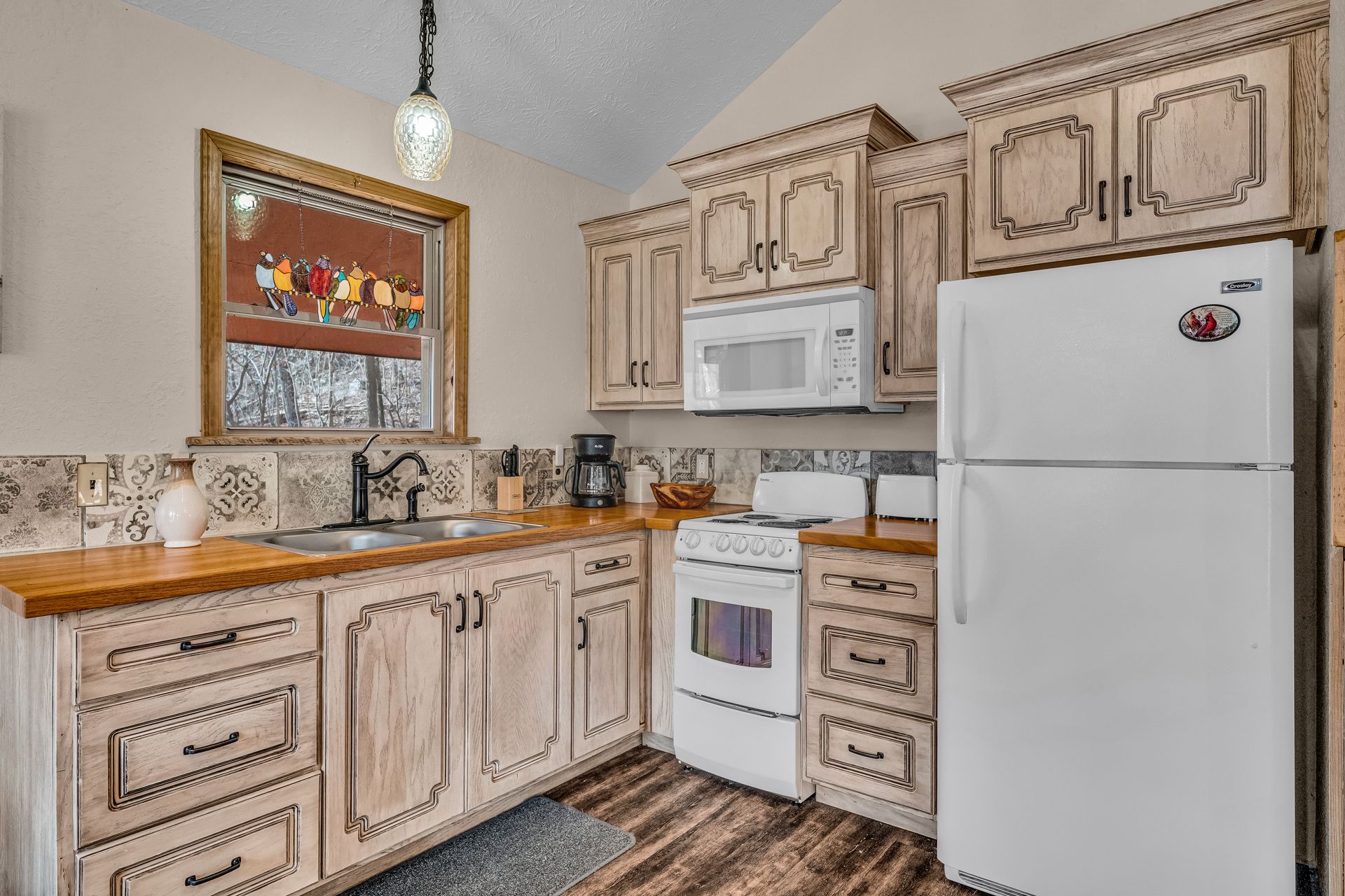 Bright kitchen with light wood cabinets, white appliances, patterned rug, and a stained-glass window