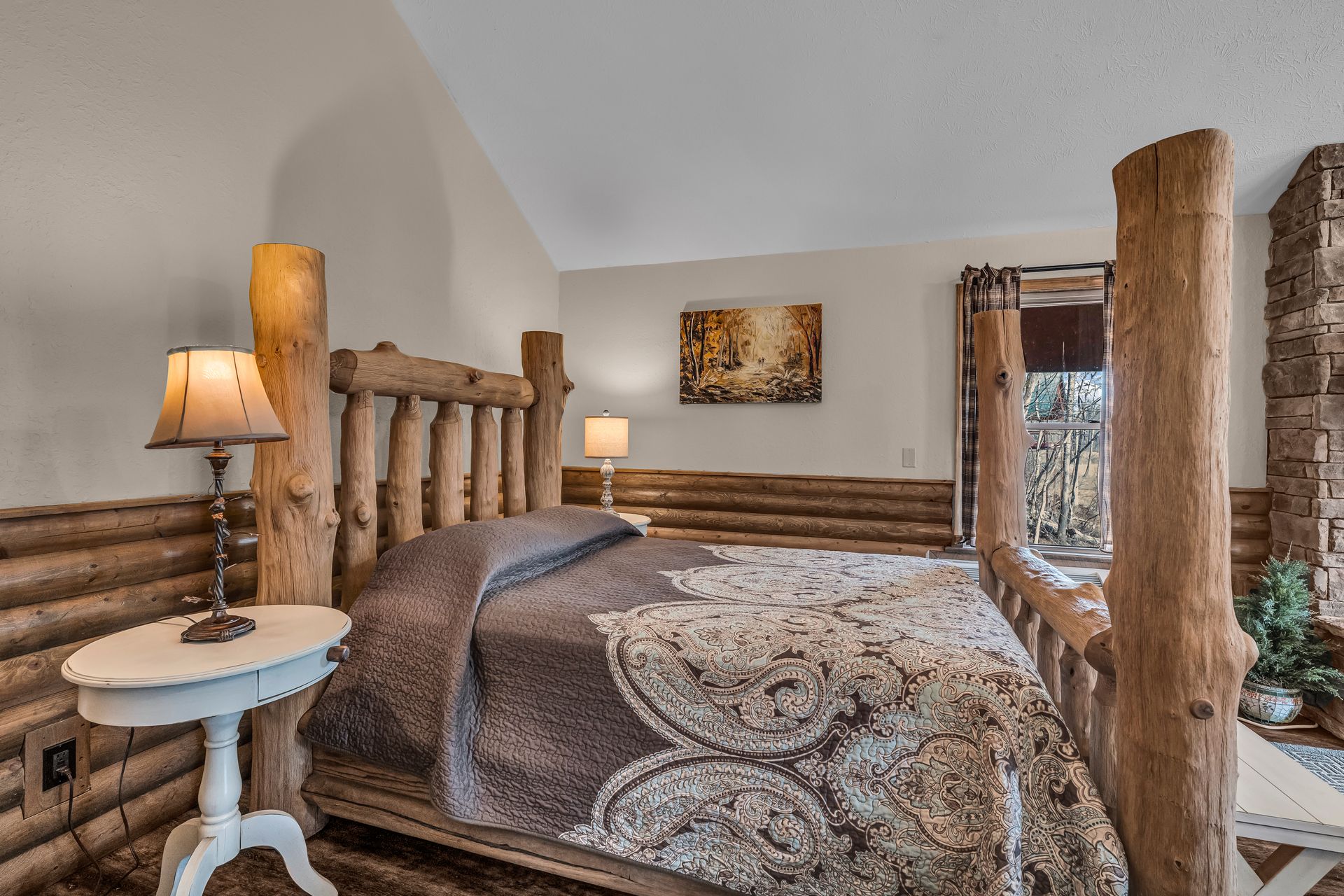 Rustic log bed with patterned quilt in a cozy cabin bedroom, with a lamp and window.