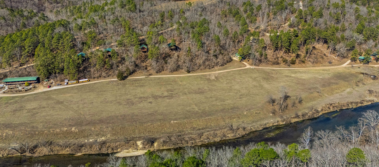 Aerial view of a grassy field beside a wooded hillside and a river, with a small green building on the left.