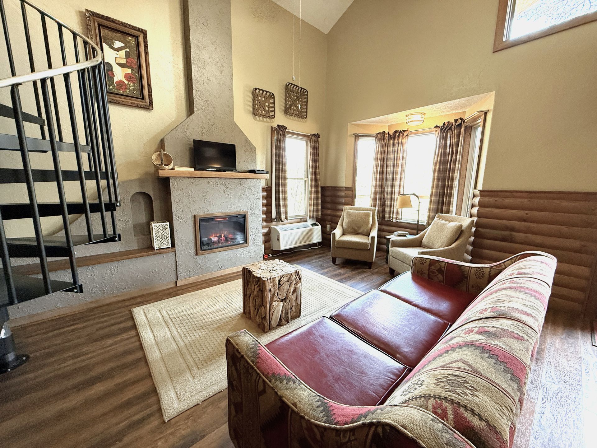 Cozy living room with spiral staircase, fireplace, armchairs, and patterned sofa by a window.