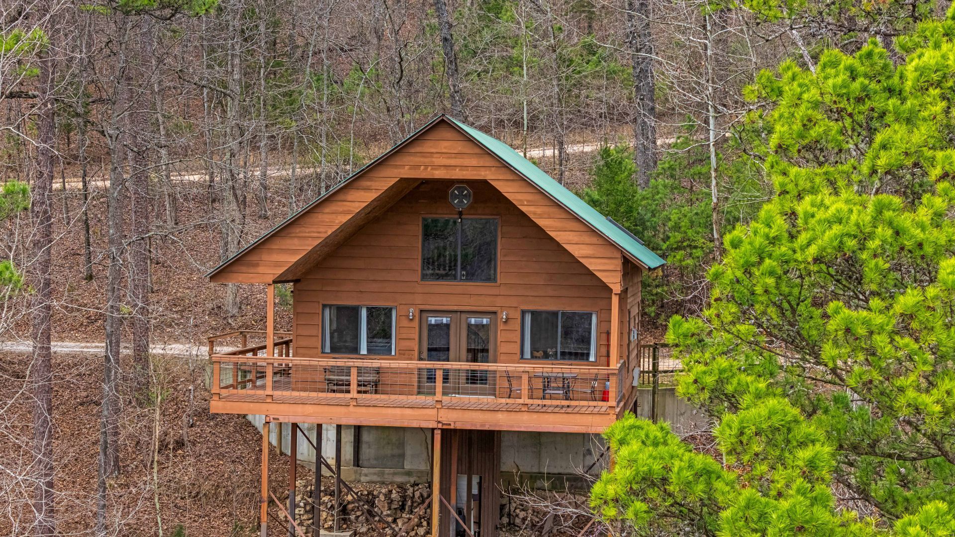 Wooden cabin with a wraparound deck on stilts in a wooded hillside setting