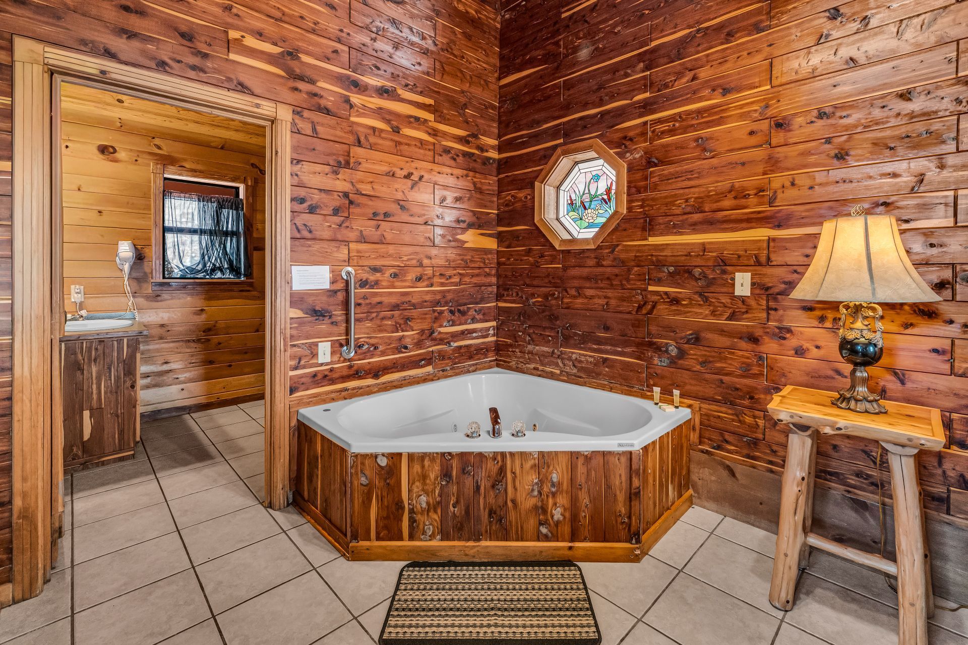 Wood-paneled bathroom with a corner soaking tub, tiled floor, lamp, and adjacent vanity area