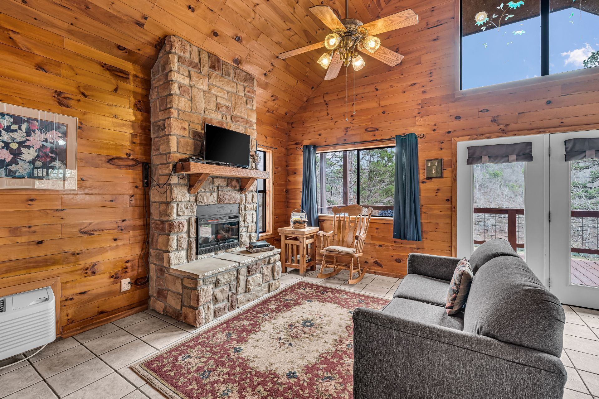 Cozy cabin living room with stone fireplace, wood walls, gray sofa, patterned rug, and ceiling fan