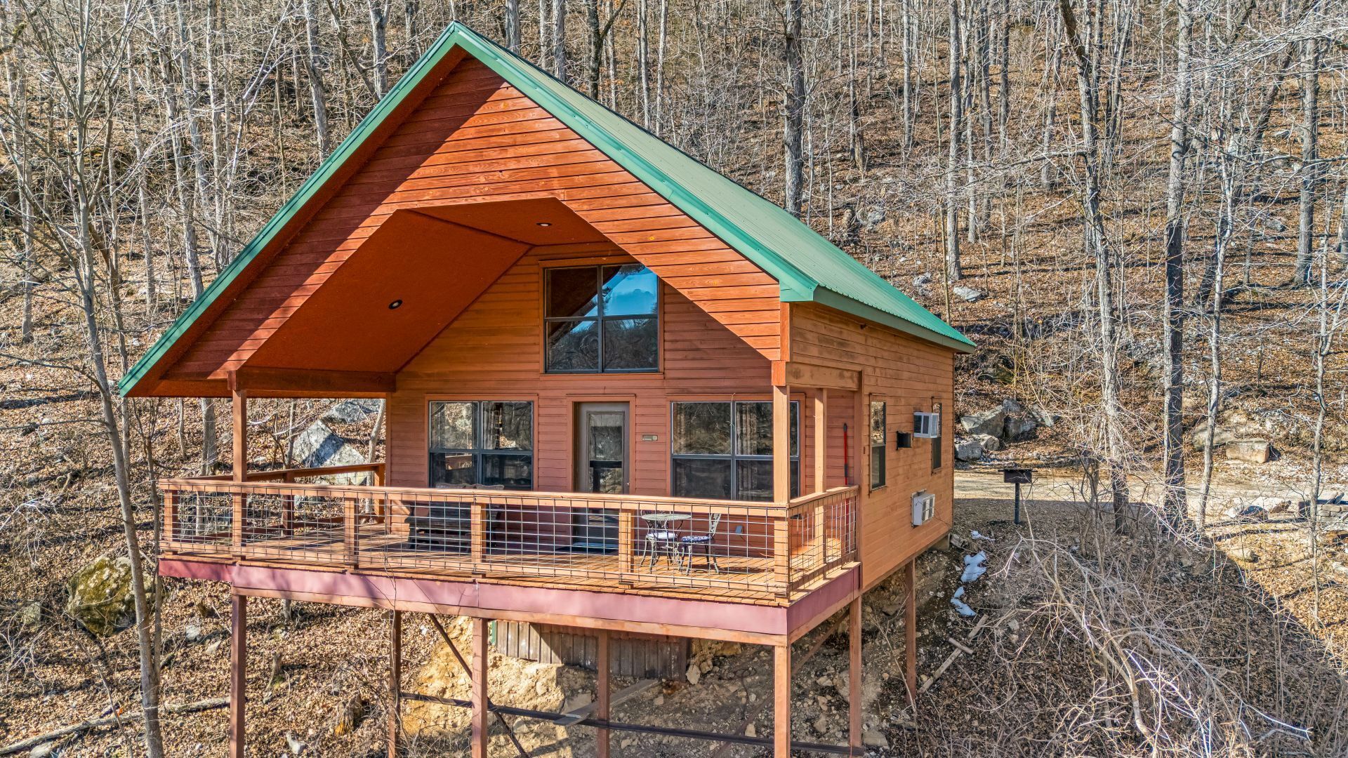 Rustic cabin with a green roof and wraparound deck in a leafless wooded hillside.
