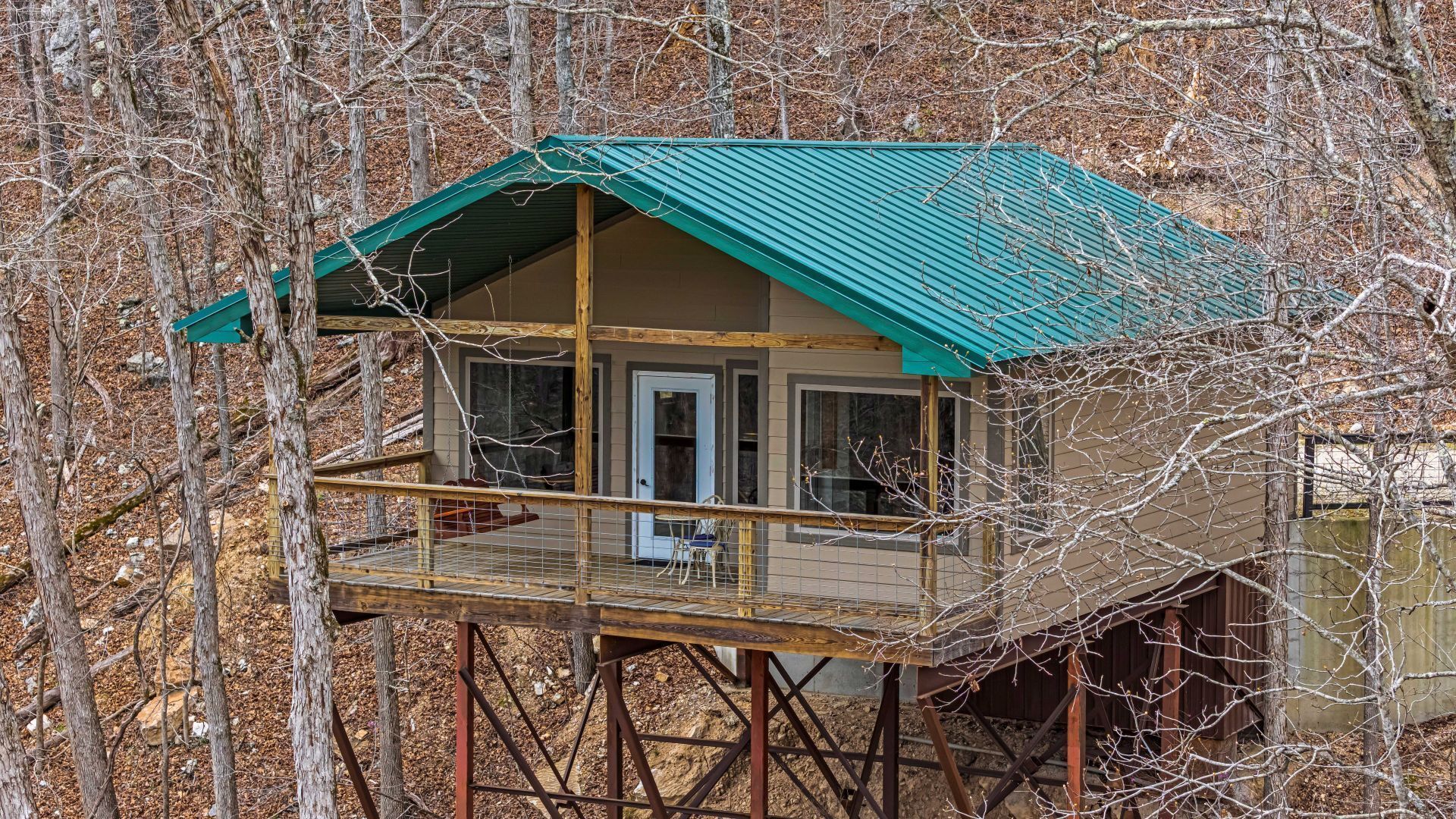 Small cabin with a teal roof and wraparound deck on stilts in a wooded area