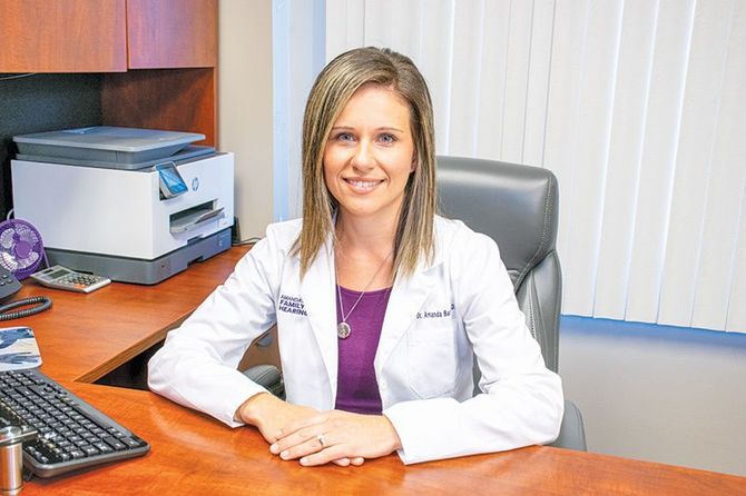 Woman in lab coat sitting at desk, smiling. Office setting with printer and computer.