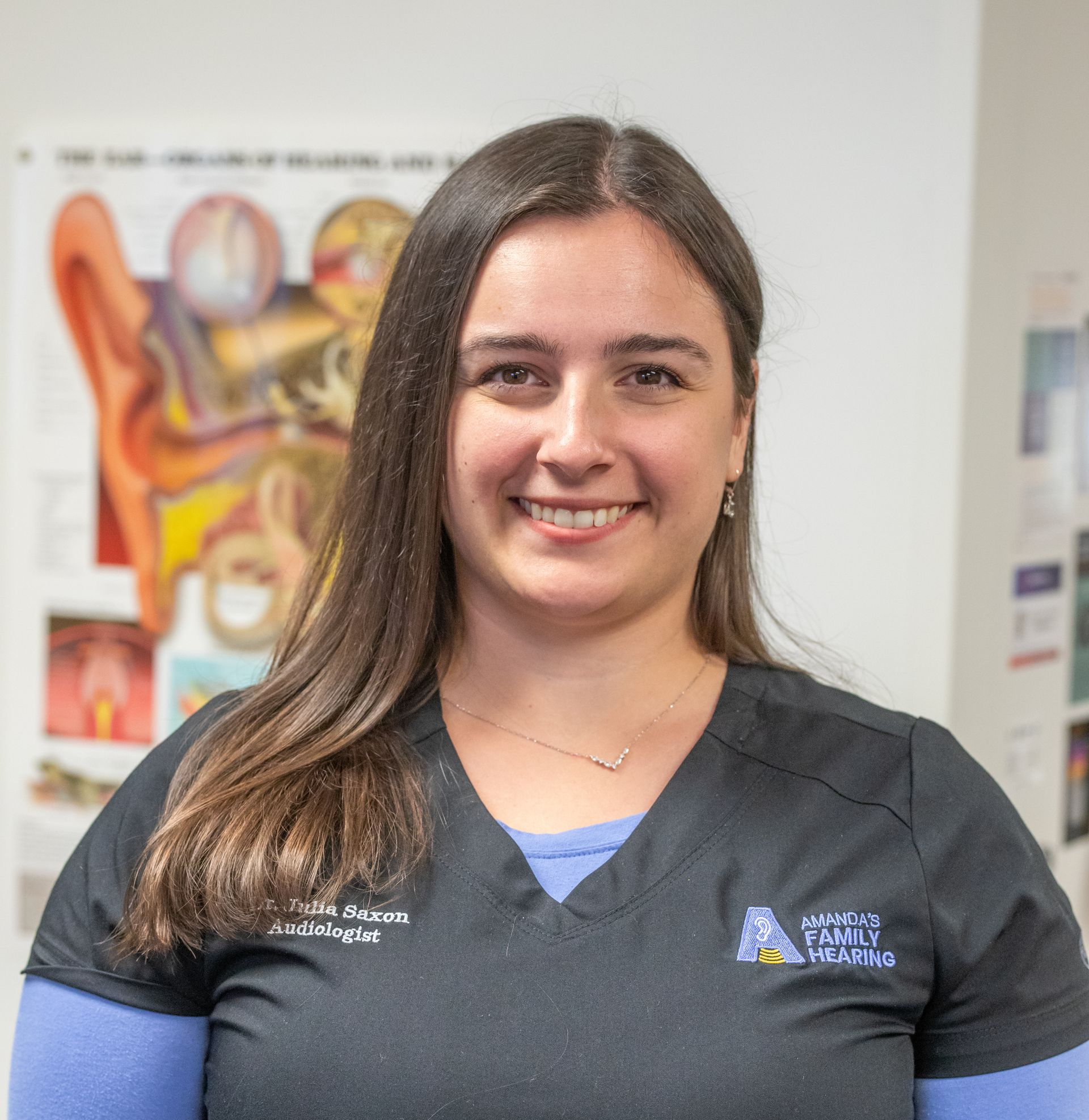Woman smiling in scrubs, standing in front of ear anatomy chart, logo on shirt.