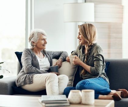 Woman and older woman on a couch, holding mugs and talking in a living room.