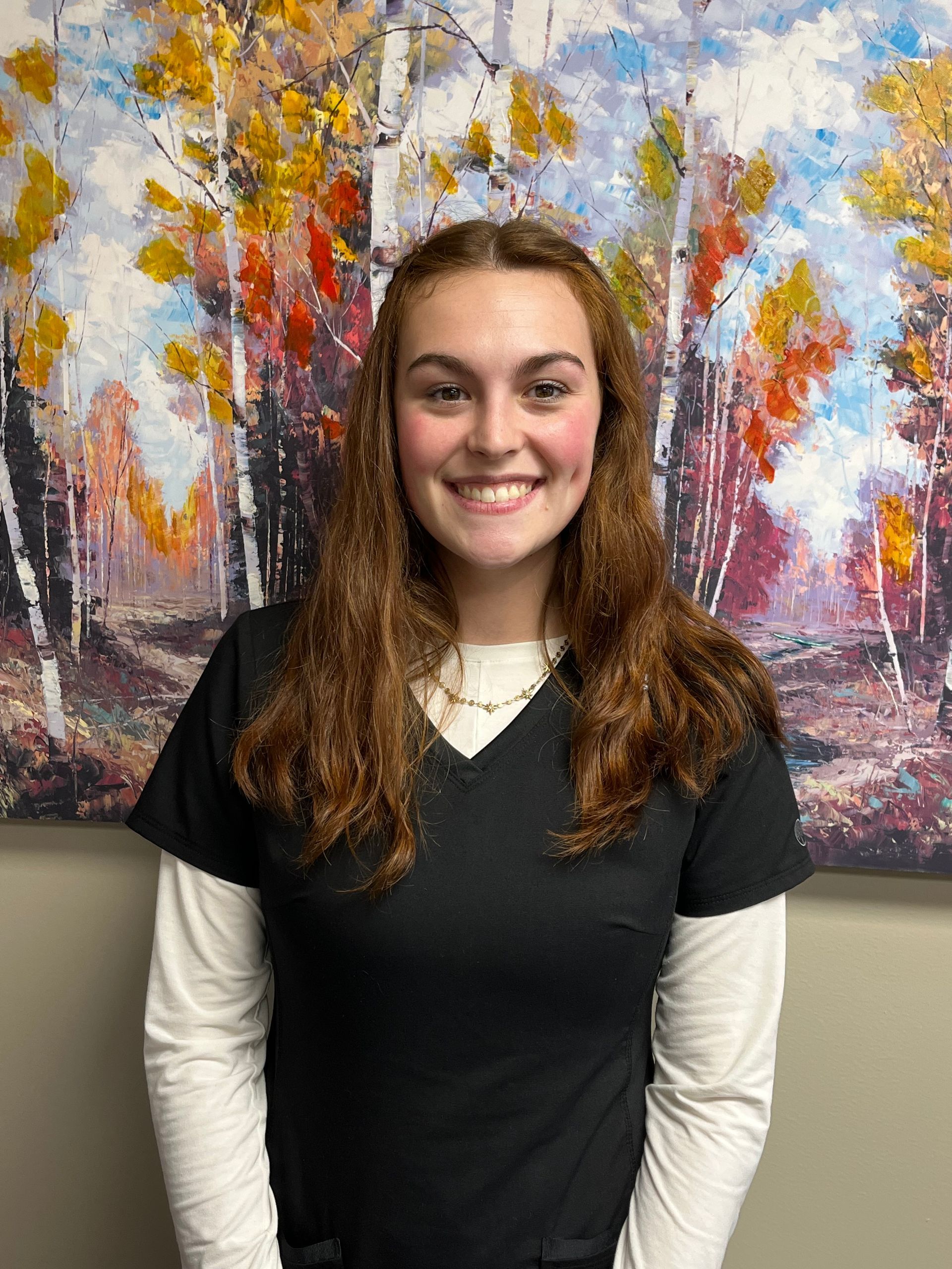 Woman smiling in black scrubs over a white shirt, with a colorful abstract painting background.