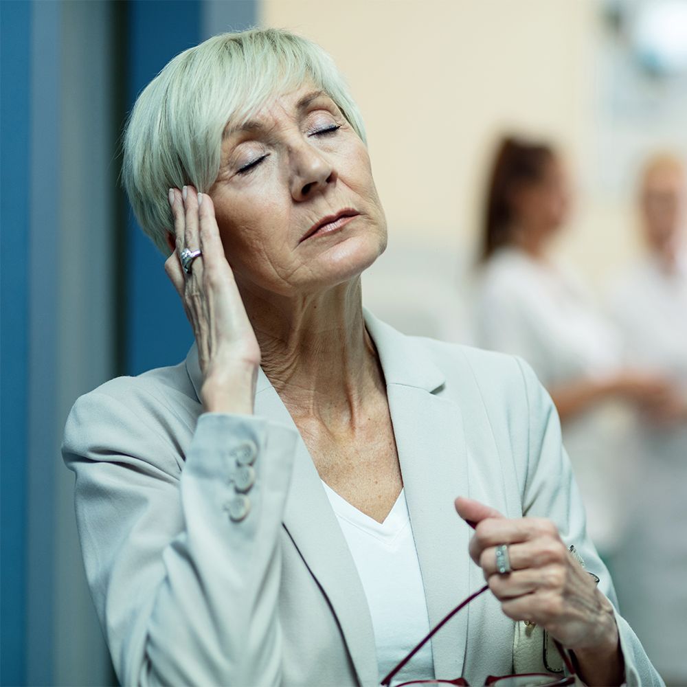Woman holding head, eyes closed; likely in pain. Wearing blazer, ring; blurred background.