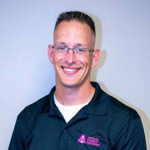 Man with glasses smiling, wearing a black shirt with a company logo, in front of a plain background.