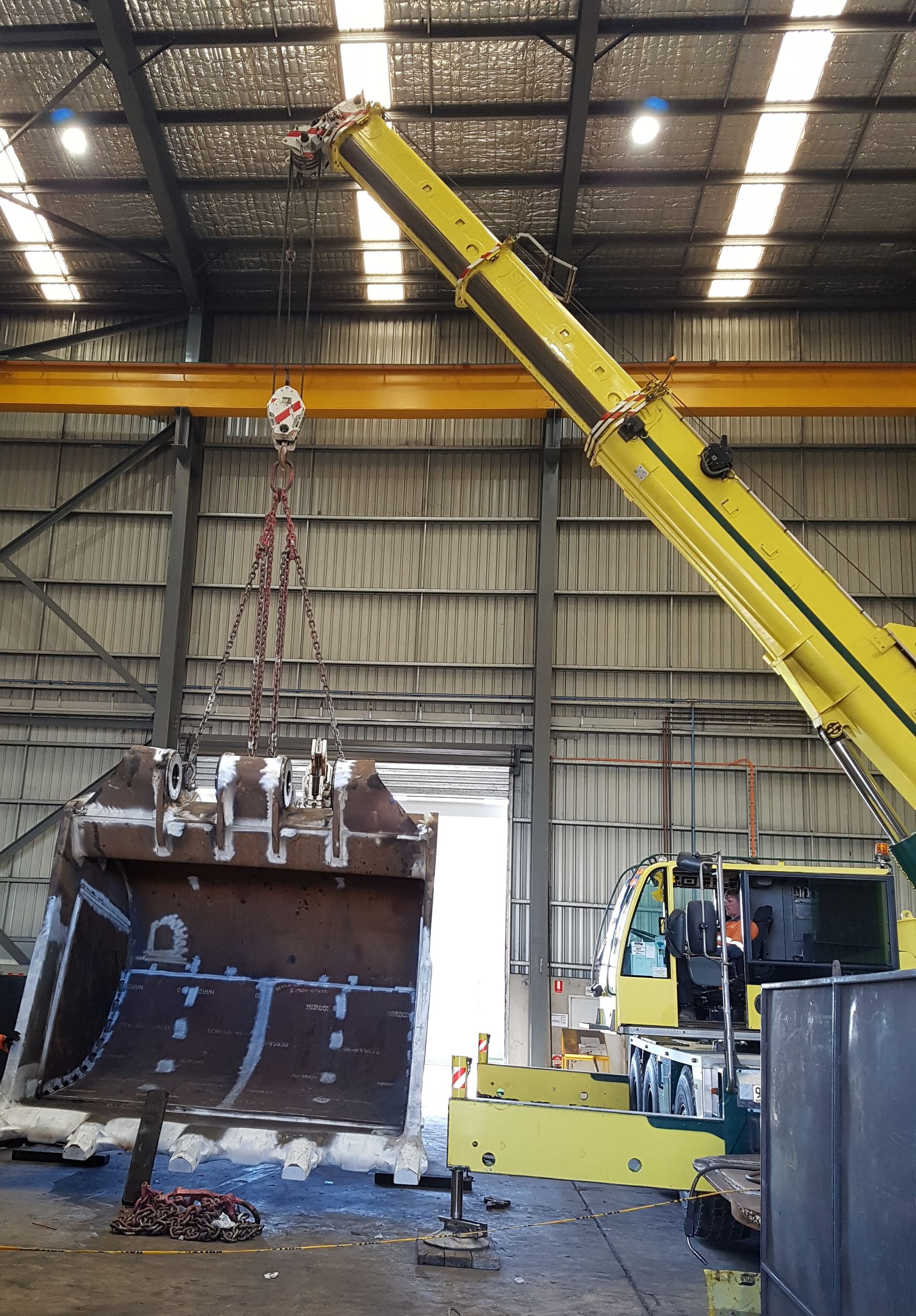 A yellow crane lifting a large metal excavator bucket inside a warehouse — Statewide Lift & Shift Pty Ltd In Paget, QLD