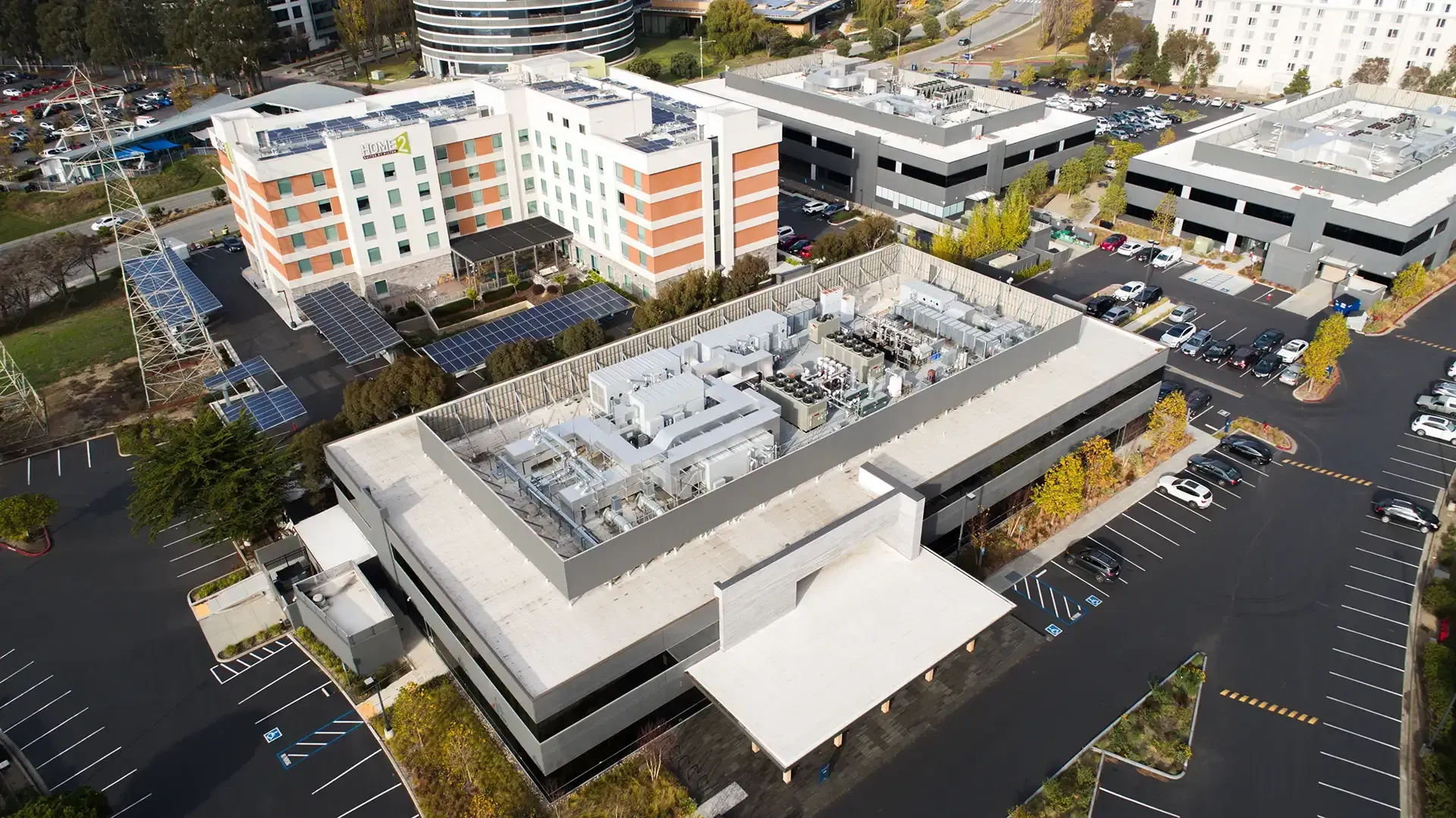 Aerial view of a commercial complex featuring a multi-story hotel, an office building with rooftop equipment, and parking.
