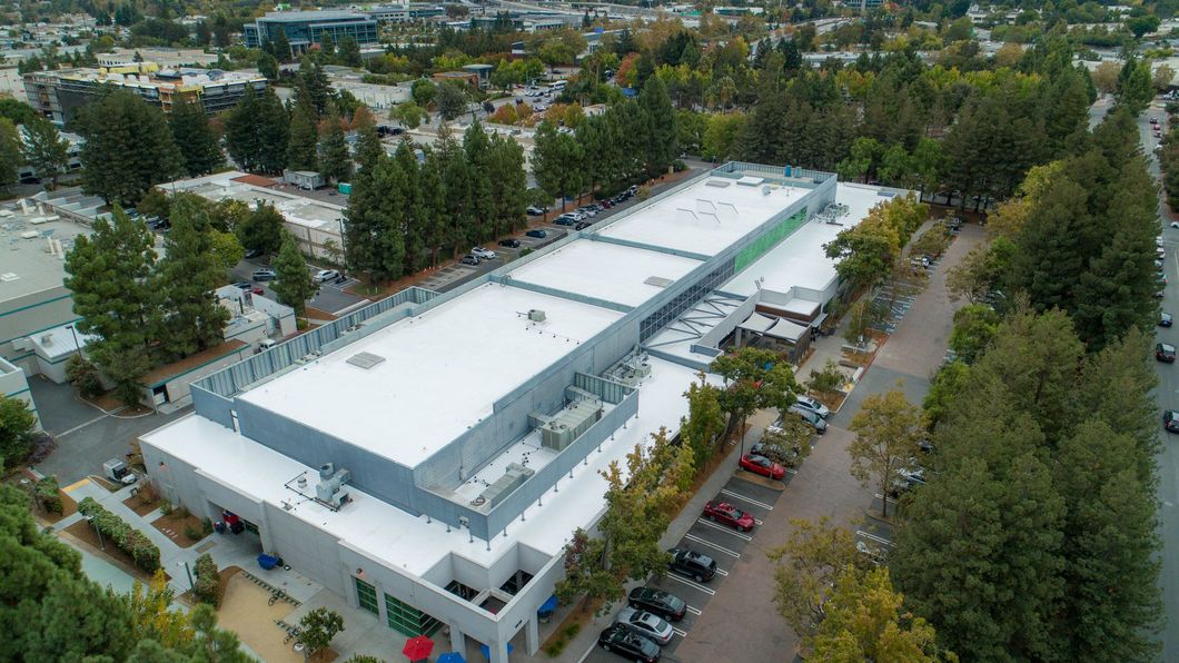 An aerial view of a flat-roofed building featuring several rows of solar panels, surrounded by trees and a parking lot.