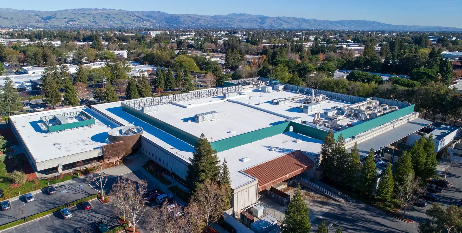 Aerial view of a large commercial building with a white flat roof and green trim, surrounded by parking and trees.