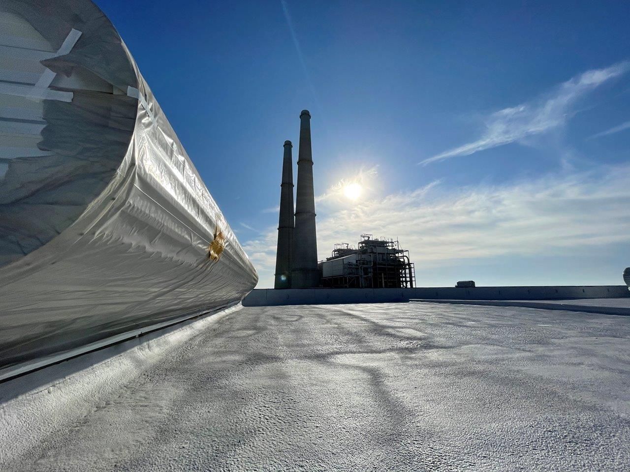 Low-angle view of a flat, textured rooftop with a industrial smokestack complex in the distance under a sunny blue sky.