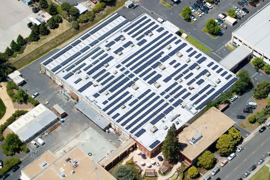 Aerial view of a commercial building with a large rooftop solar panel array surrounded by parking lots and landscaping.