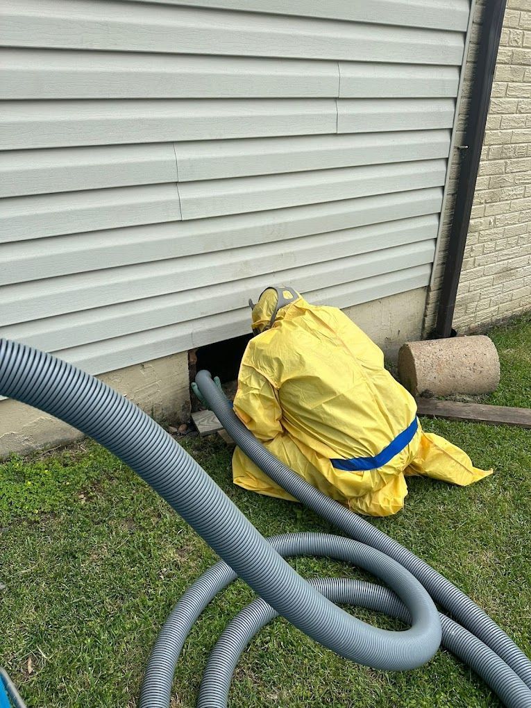 A person in a yellow hazmat suit kneeling by a foundation vent, inserting a large gray hose into the opening.