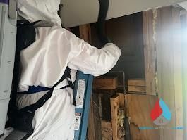 A person in a white hazmat suit and backpack vacuum cleans debris from inside an open wall cavity.