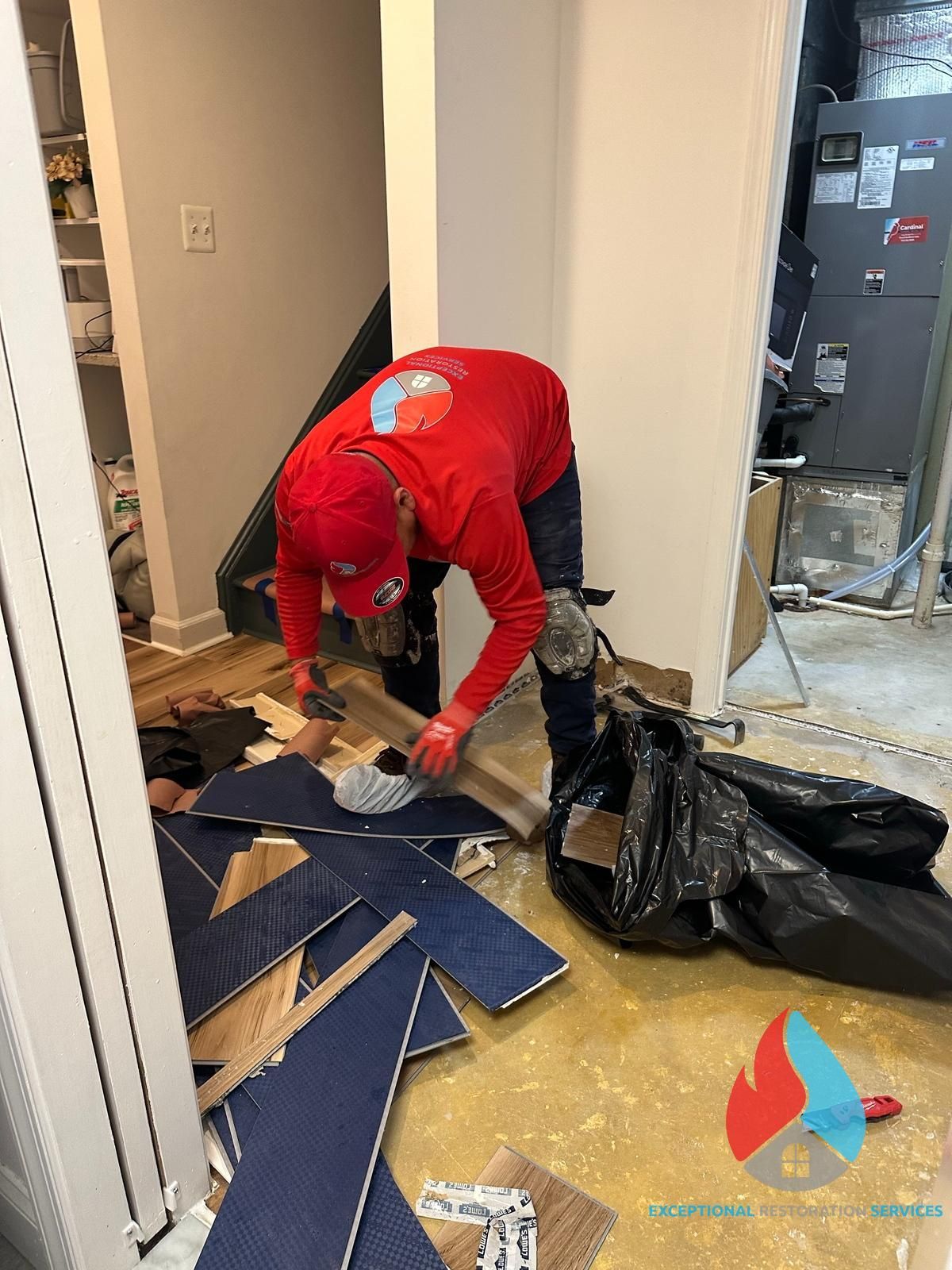 A worker in a red shirt removing flooring debris and placing it into a black trash bag in a residential room.