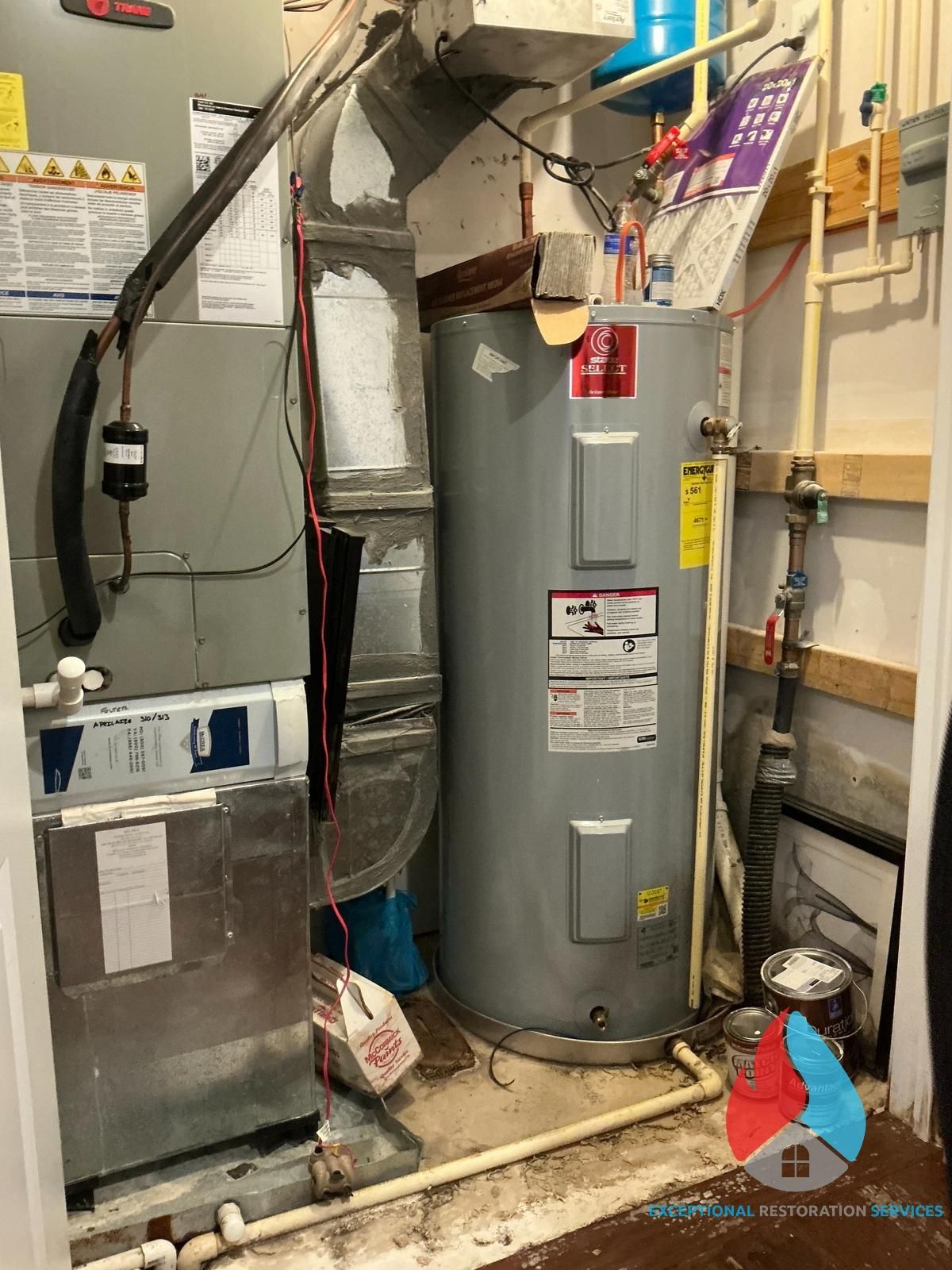 Mechanical closet with a gray water heater tank positioned beside a large Trane HVAC unit, pipes, and ventilation ducts.