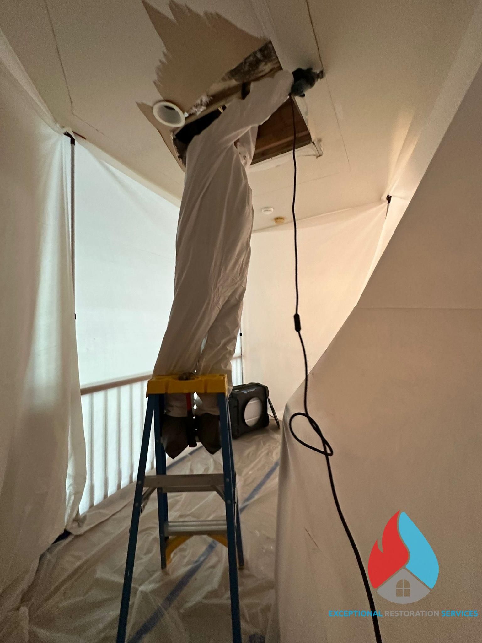 A worker in a white protective suit on a ladder cuts a rectangular opening in a ceiling covered by protective plastic.