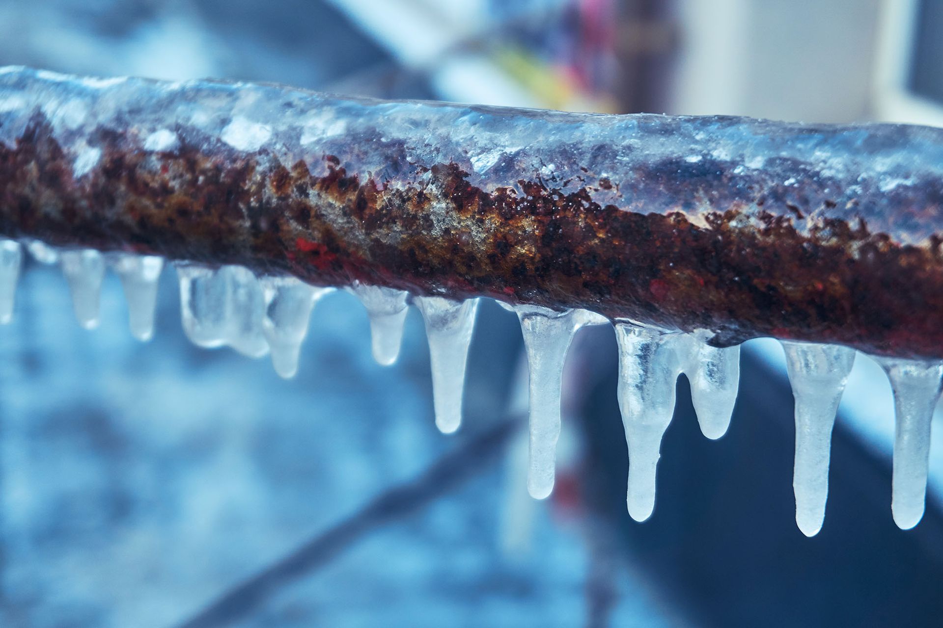 A rusted, horizontal metal bar covered in ice with icicles hanging from the bottom edge against a blurred, cold background.