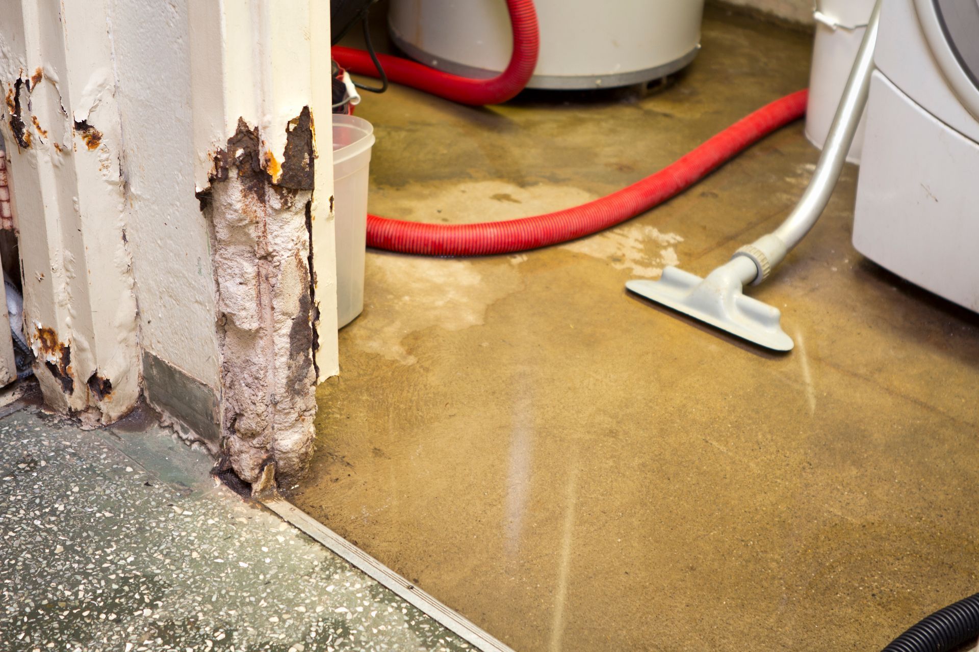 A vacuum cleaner nozzle sits on a wet concrete floor near a damaged, water-stained door frame and red hoses.