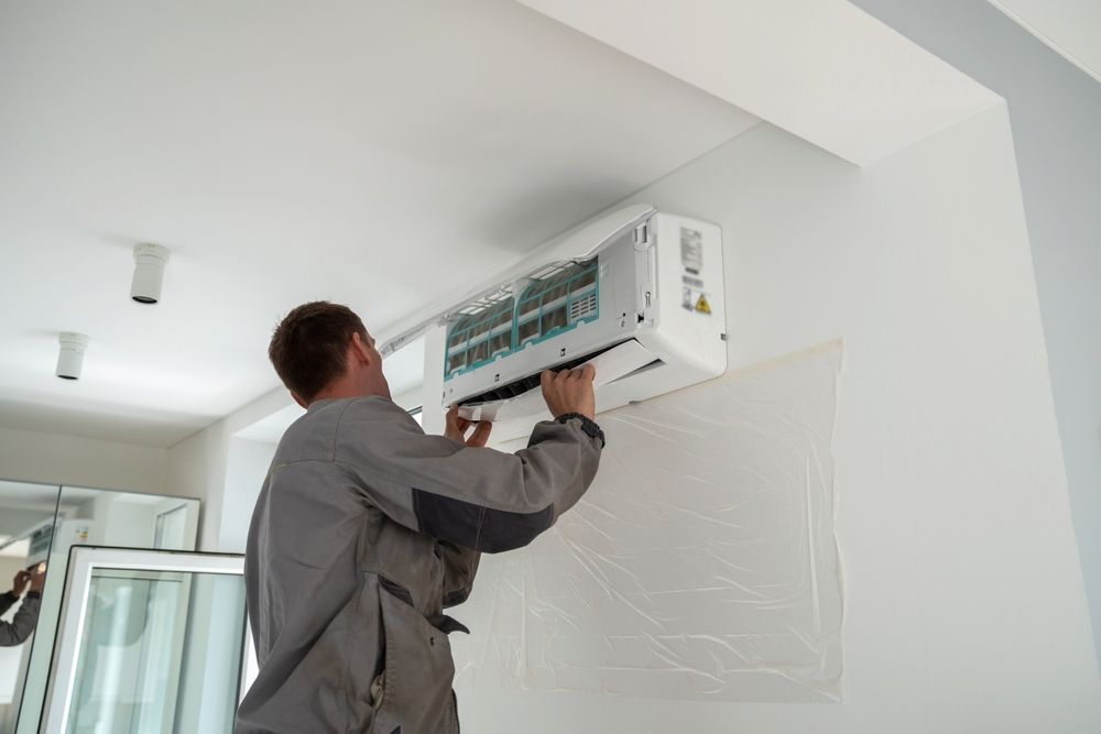 Man in Gray Jumpsuit Installing a White Air Conditioner on a White Wall Indoors — White Rose Electrical in Coffs Harbour, NSW