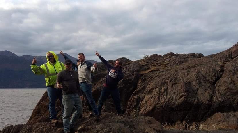Four men pose jauntily on rocky shore with mountains and cloudy sky in background.
