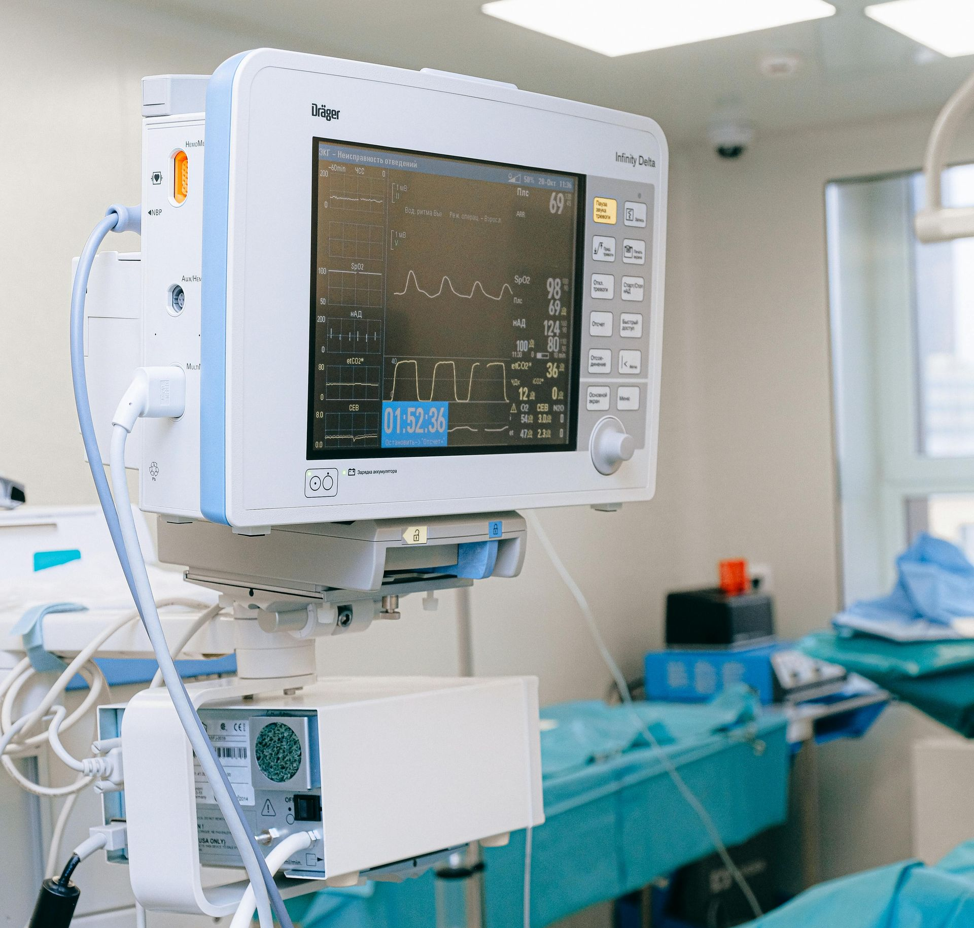Hospital patient monitor beside a bed in a brightly lit operating room, with a patient draped in blue sheets.