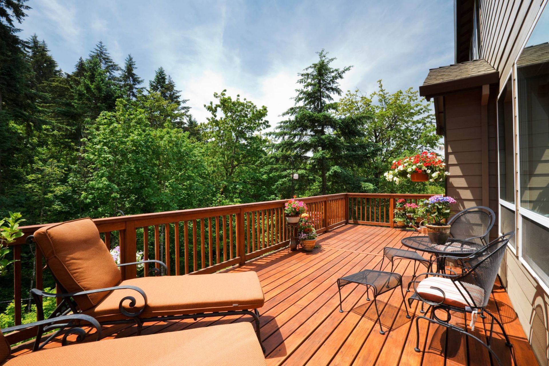 Wooden deck with lounge chair, table, and chairs. Overlooks a lush forest on a sunny day.